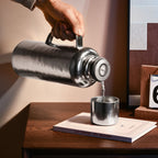 Pure titanium 1.2L thermal carafe pouring hot water into cup, temperature dial on lid, sunlit desk scene