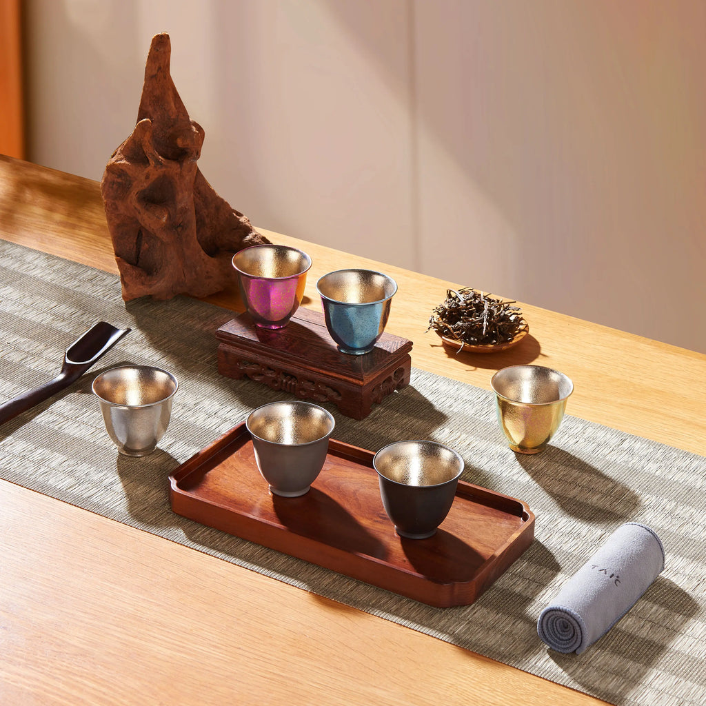 Titanium double-wall tea cups arranged on woven runner with wooden tray, driftwood and loose tea leaves in sunlight