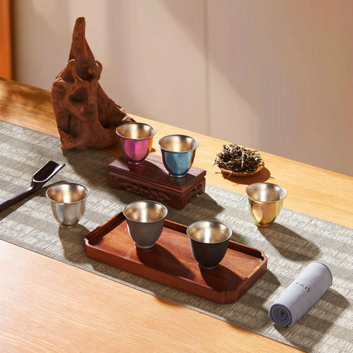 Titanium double-wall tea cups arranged on woven runner with wooden tray, driftwood and loose tea leaves in sunlight