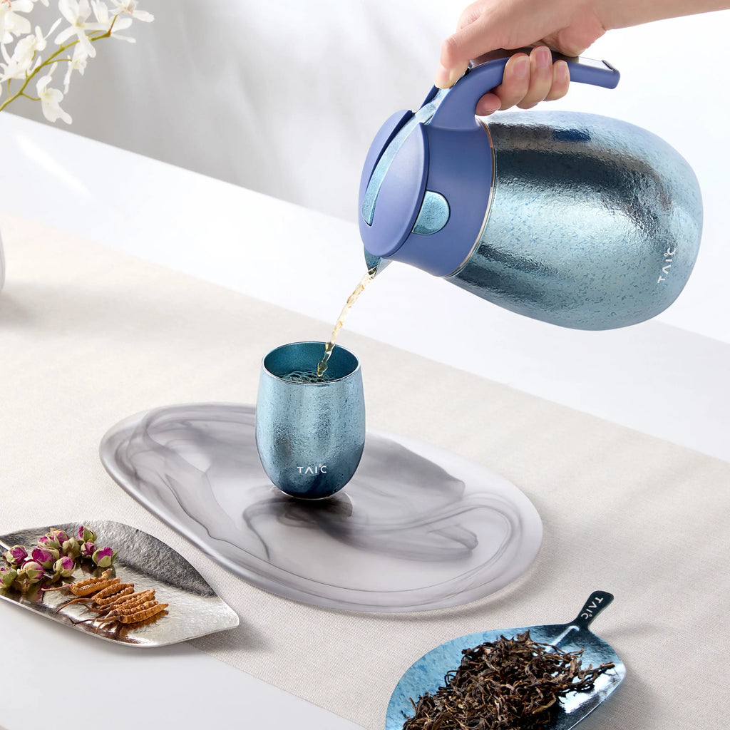 Pouring hot tea from a blue titanium thermal carafe into a matching titanium cup on a marble tray