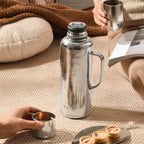 Pure titanium thermal carafe uncapped on cozy rug with hands holding tea cups and dessert plate nearby