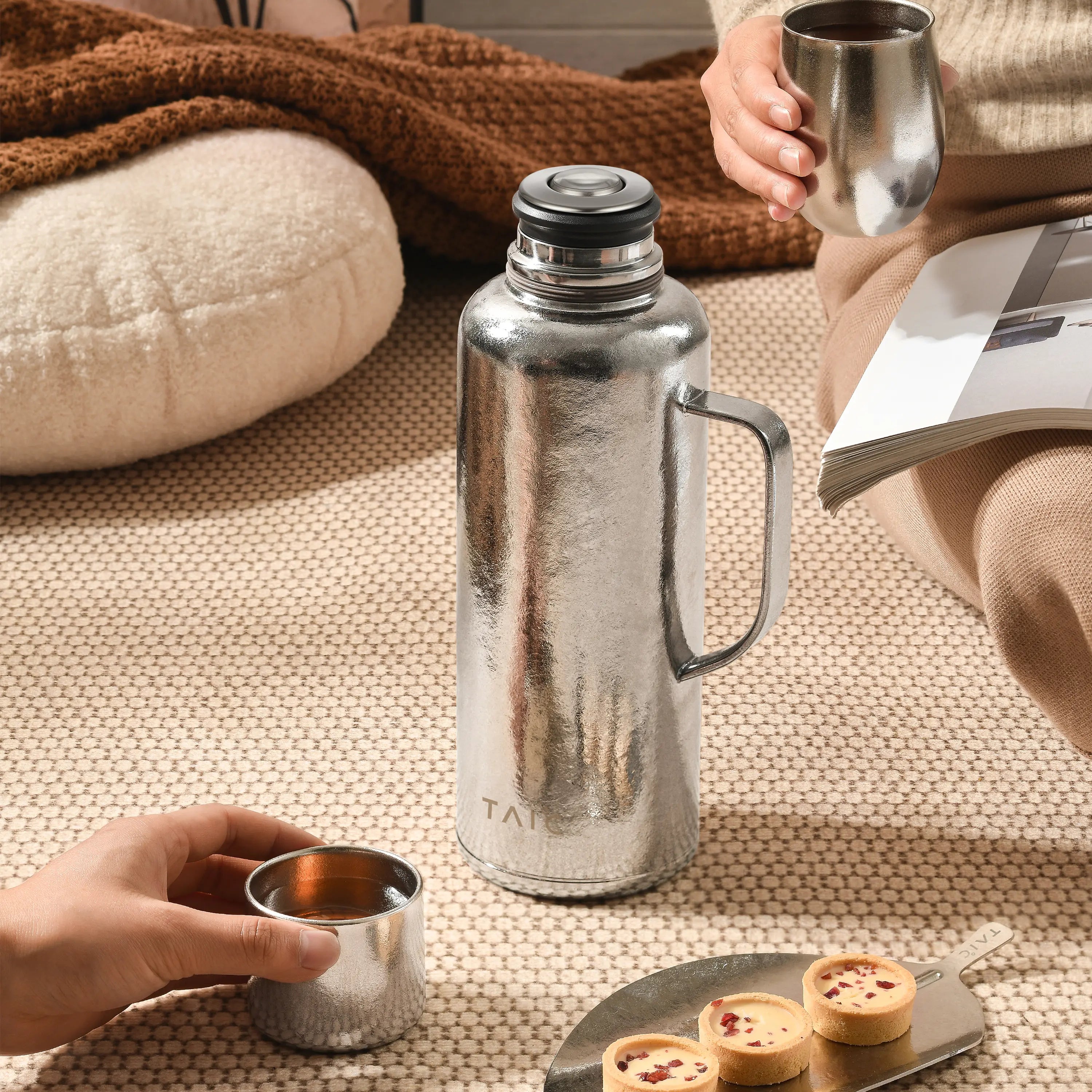 Pure titanium thermal carafe uncapped on cozy rug with hands holding tea cups and dessert plate nearby