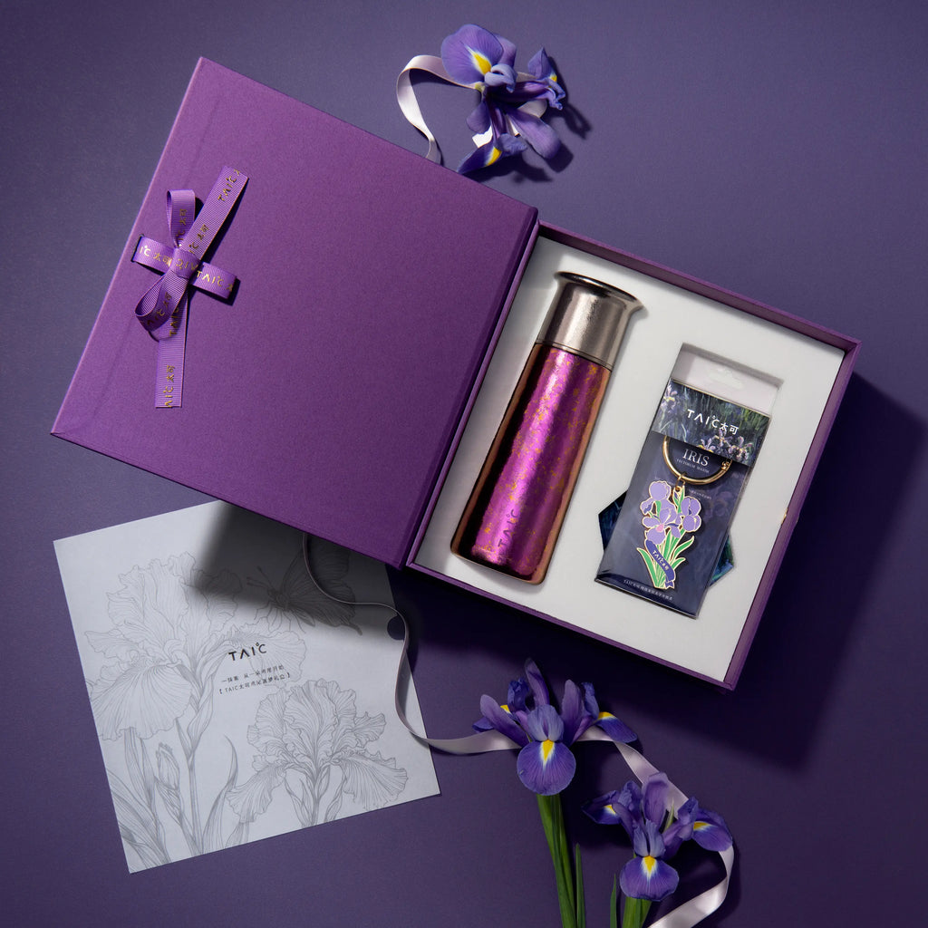 Top-down flat lay of open titanium tumbler gift box with iris charm, sketch card and fresh purple iris blooms