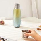 Green-to-blue Aurora titanium tea infuser tumbler on desk beside hand holding titanium cup and rose tea jar