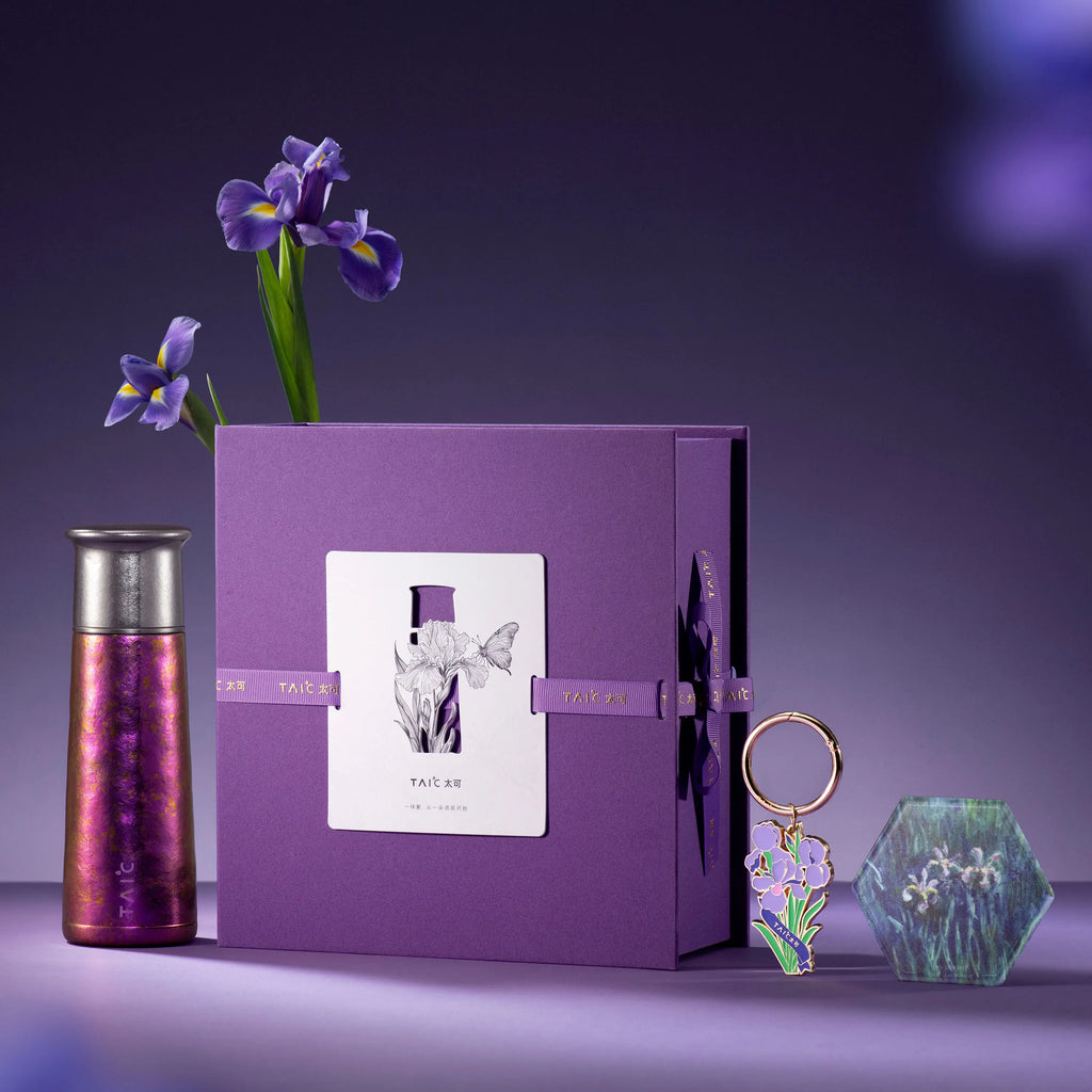Iridescent titanium tumbler standing beside closed purple gift box with iris keyring, coaster and iris flowers