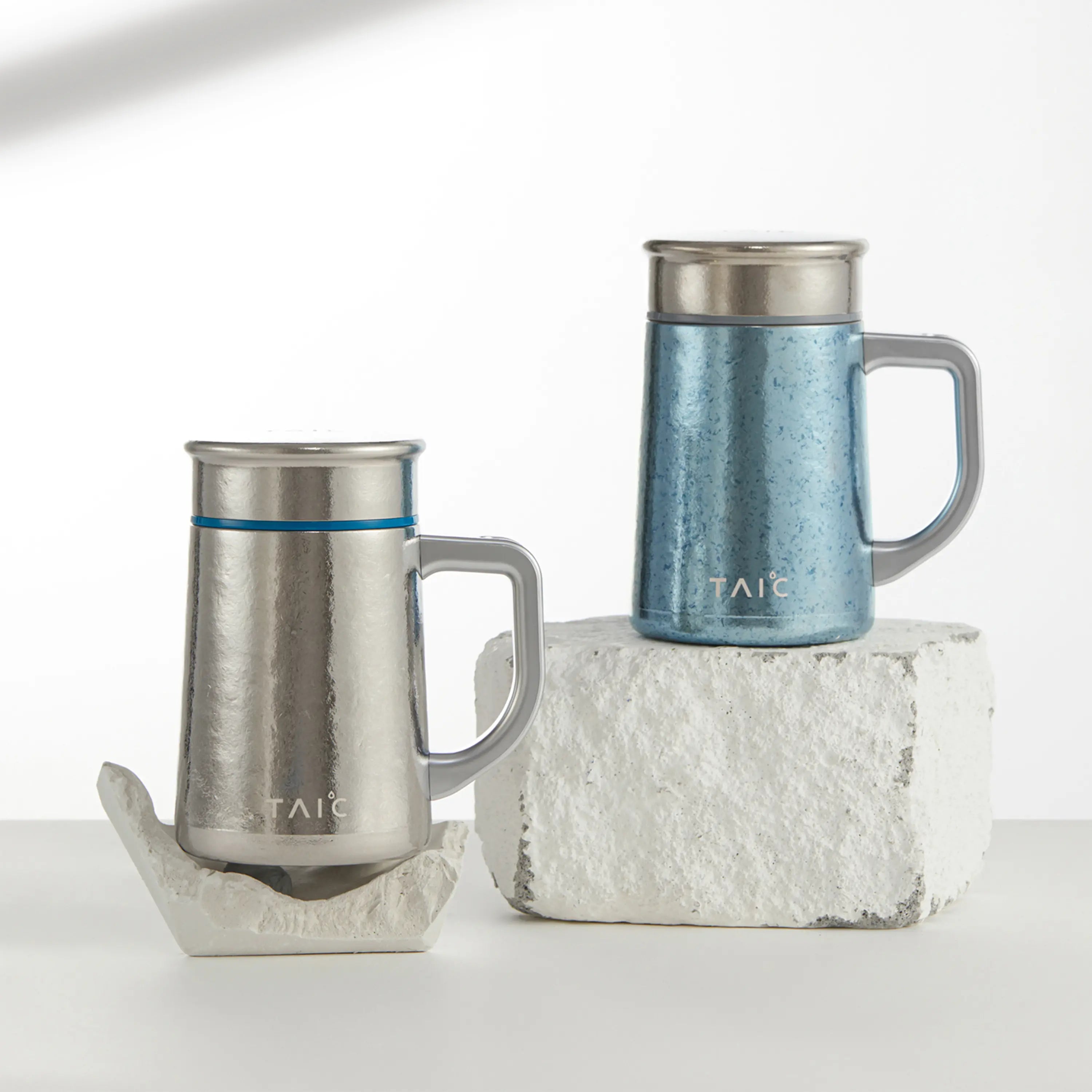 Pure titanium office mugs in silver and blue aurora finishes styled on stone blocks, lifestyle shot