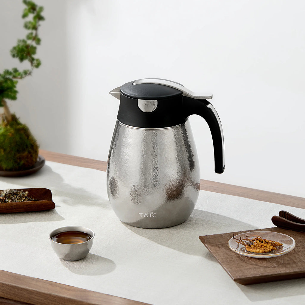 Hammered silver titanium thermal carafe on a wood tea table with a filled titanium cup and loose tea