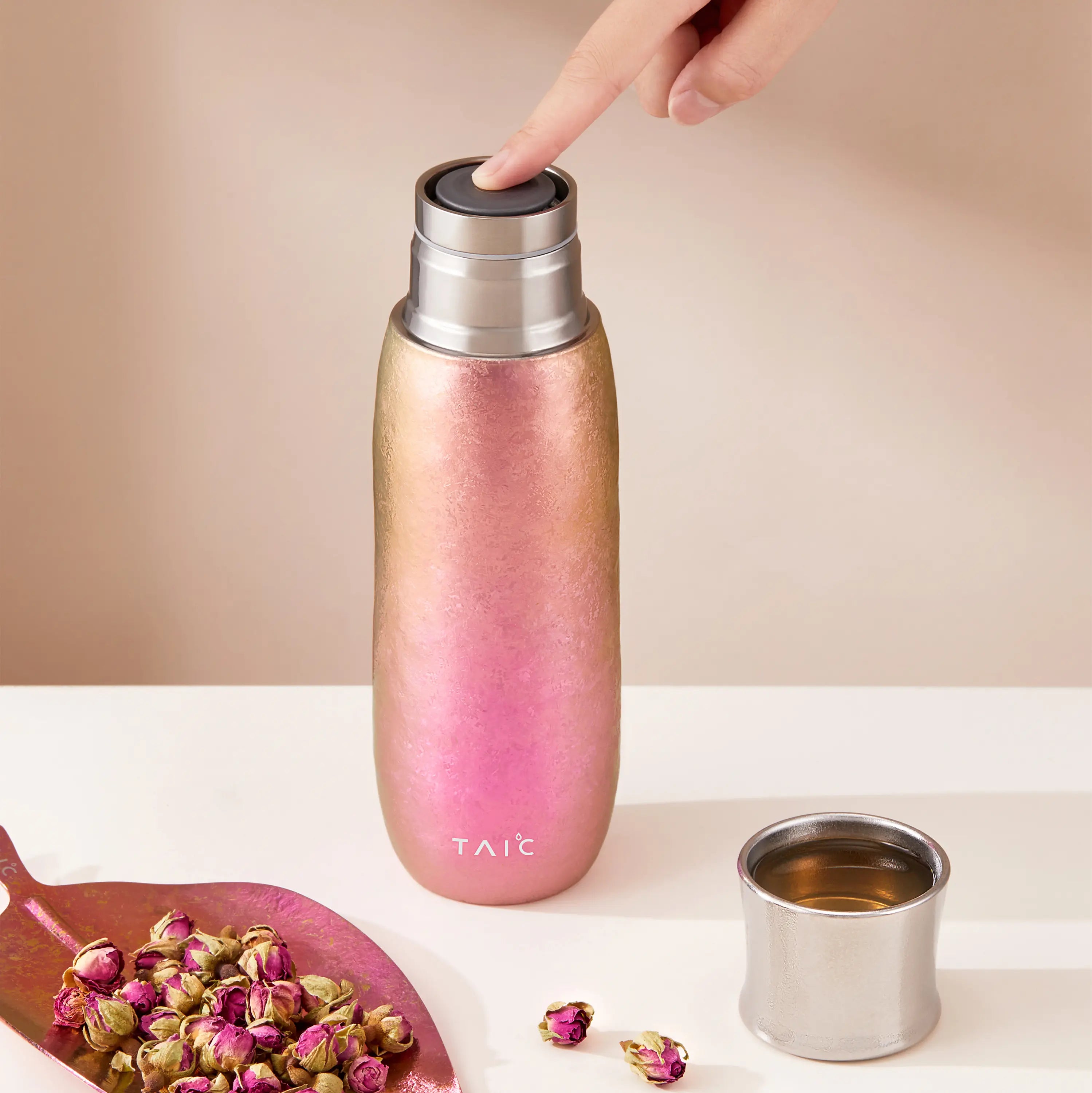 Finger pressing the push-button plug of rose-gold Aurora titanium tea infuser tumbler, dried rosebuds nearby
