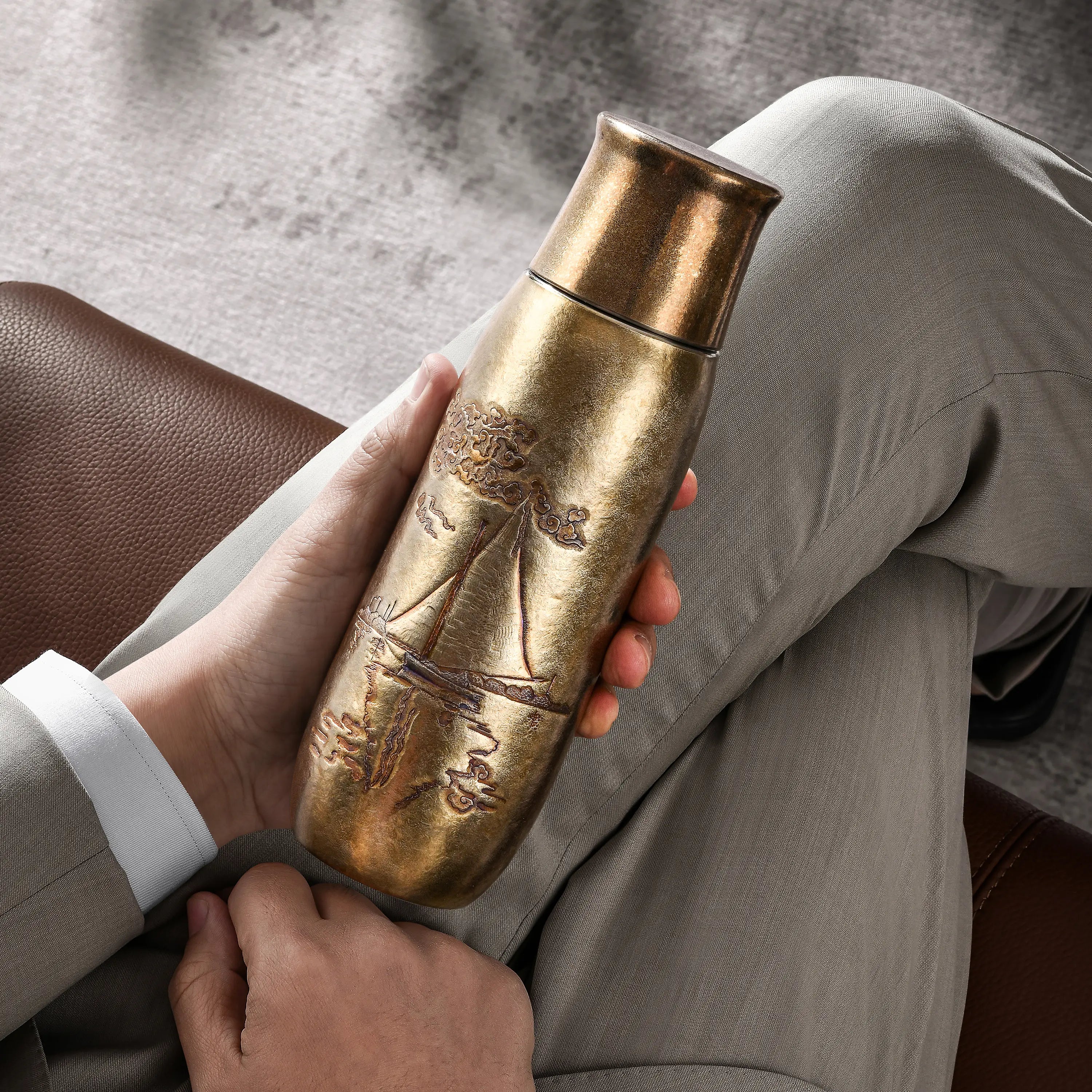 Monet Sailboat titanium infuser tumbler in golden bronze finish, held on the lap by a seated man, lifestyle shot
