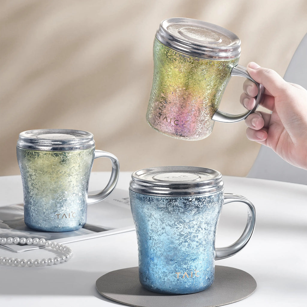 Three titanium mugs in green, blue and rainbow finishes with lids, one lifted by hand