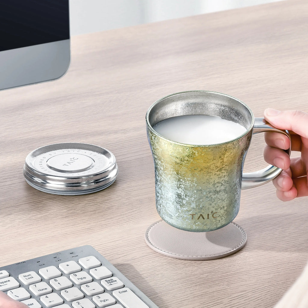 Titanium insulated mug filled with milk, held by hand on a desk beside keyboard and open lid
