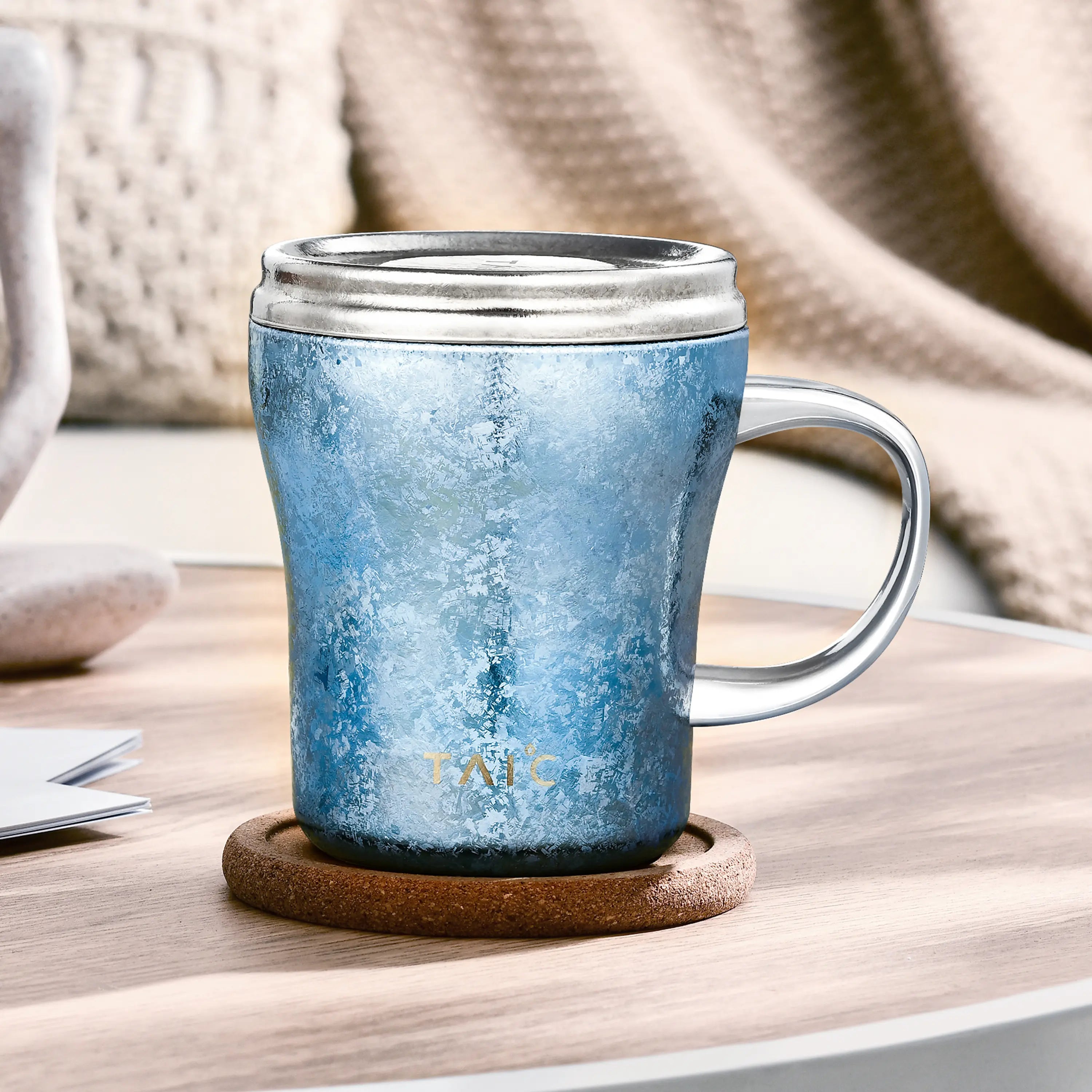 Frosted blue titanium mug with lid on a cork coaster in a cozy knit-blanket setting