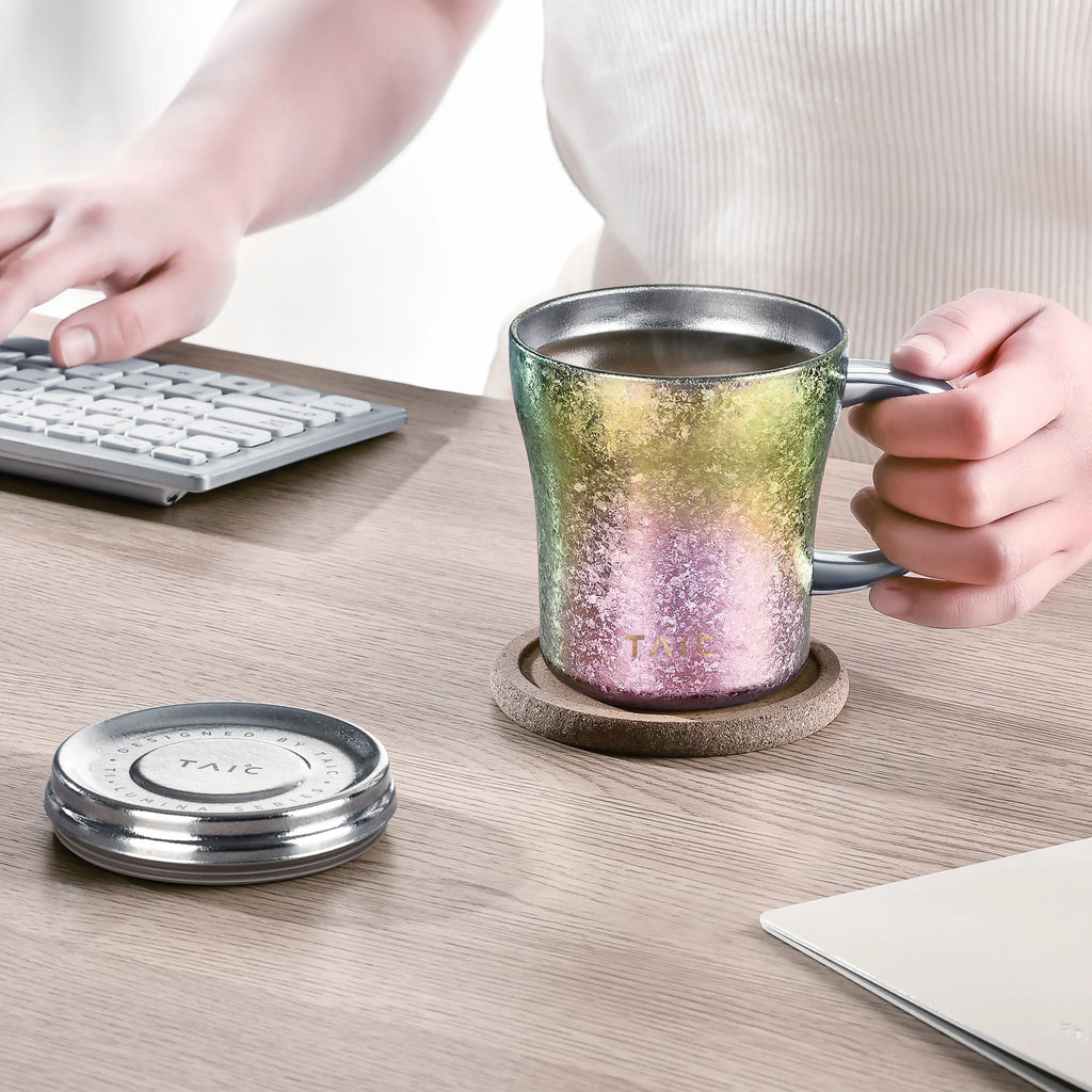 Iridescent green-to-pink titanium mug on a coaster at a keyboard desk, lid set aside