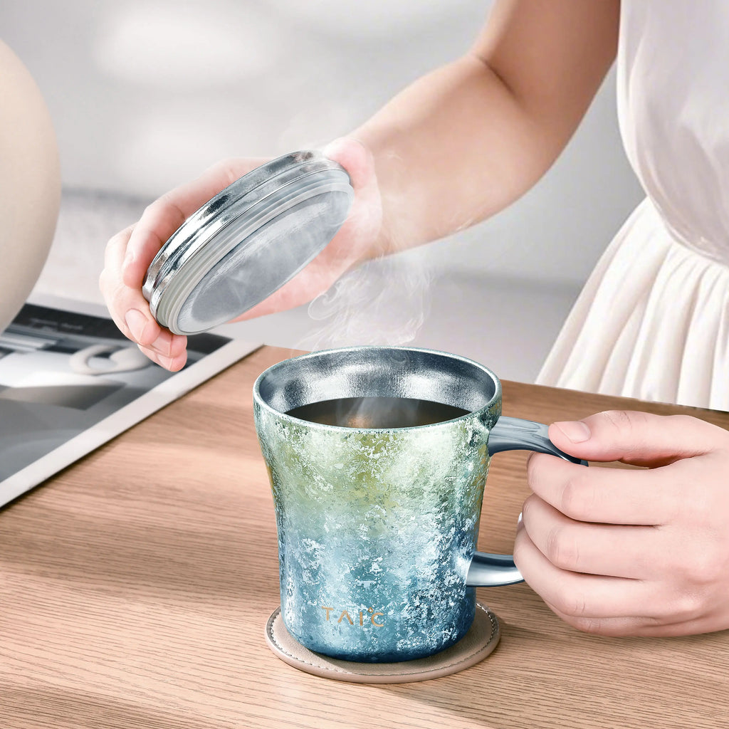 Titanium coffee mug with steaming brew as a hand lifts its lid, lifestyle desk scene