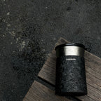 Кофейная кружка BlackInTi из чистого титана — кружка лежит на состаренной деревянной доске над тёмным камнем